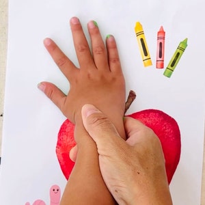 May include: A child's hand is being used to make a handprint on a red apple. The apple is on a white piece of paper with a pink worm and three crayons.