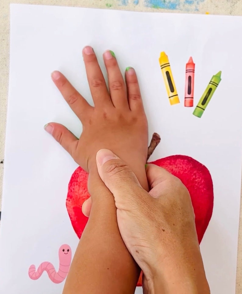 May include: A child's hand is being used to make a handprint on a red apple. The apple is on a white piece of paper with a pink worm and three crayons.