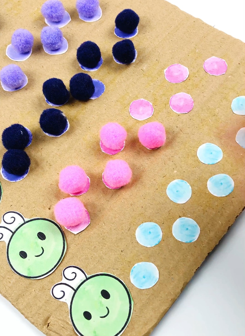 May include: A brown cardboard surface with various colored pom-poms, including pink, blue, and purple. There are also blue, pink, and light blue circles on the surface. Two green cartoon bugs with black eyes and smiles are on the bottom of the surface.