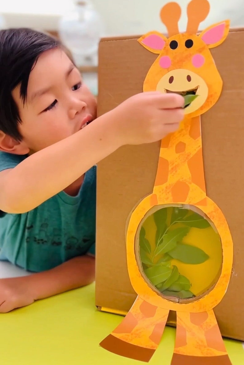 K&ouml;nnte beinhalten: Ein Kind f&uuml;ttert eine Pappgiraffe aus Pappe mit gr&uuml;nen Bl&auml;ttern. Die Giraffe hat einen braunen und orangefarbenen K&ouml;rper mit einer gro&szlig;en, runden &Ouml;ffnung f&uuml;r die Bl&auml;tter. Die Giraffe ist auf einem braunen Pappkarton montiert.
