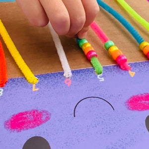 May include: A child's hand threads colorful pipe cleaners through holes in a cardboard board with a purple face drawn on it. The pipe cleaners are decorated with colorful beads.