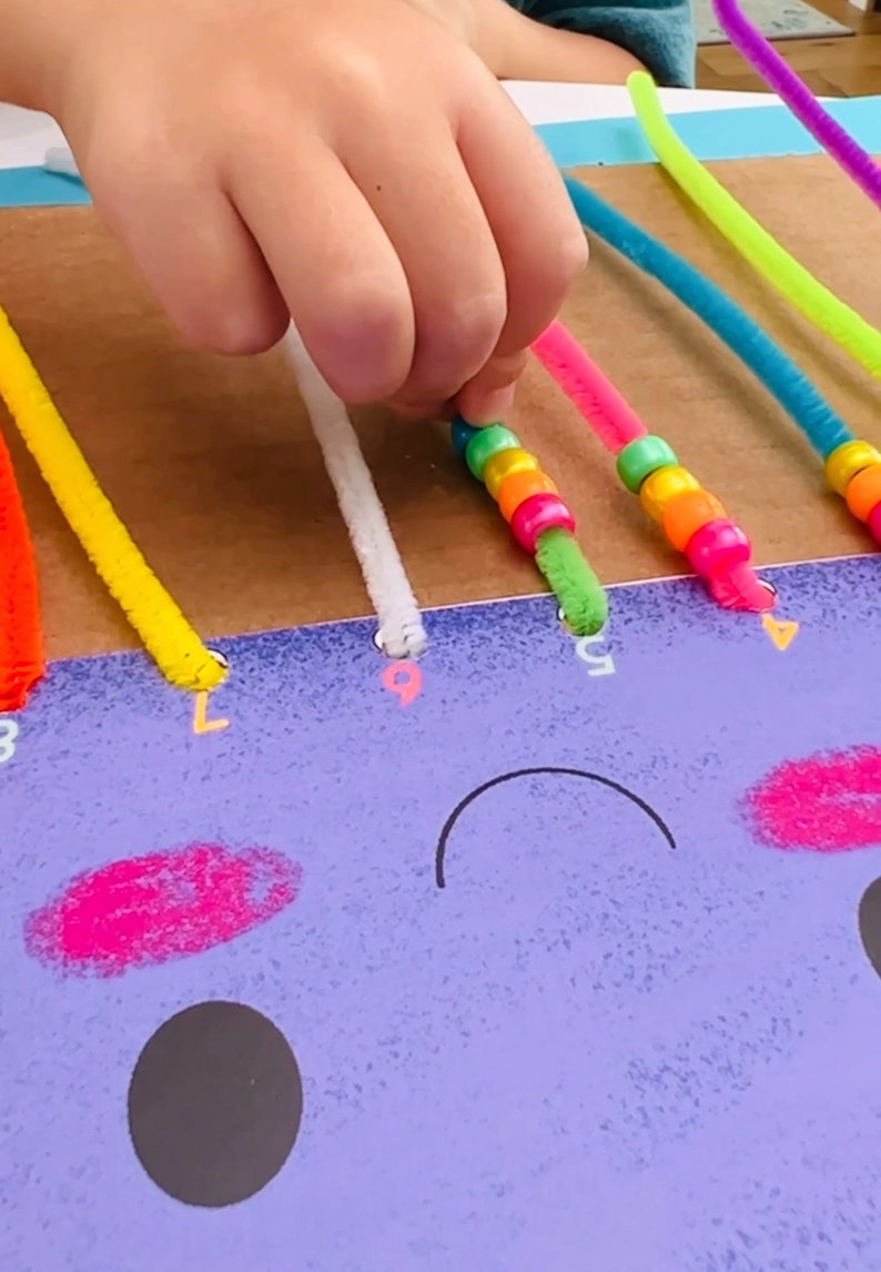 May include: A child's hand threads colorful pipe cleaners through holes in a cardboard board with a purple face drawn on it. The pipe cleaners are decorated with colorful beads.