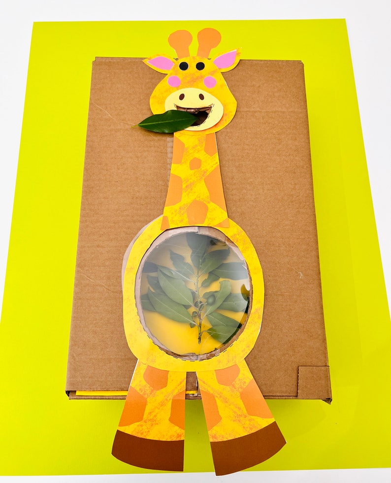 K&ouml;nnte beinhalten: Ein Karton mit einem Giraffen-Ausschnitt, der an der Vorderseite befestigt ist. Die Giraffe ist gelb und braun mit einem gr&uuml;nen Blatt im Maul. Im Bauch der Giraffe befindet sich ein transparentes Plastikfenster, durch das man eine kleine gr&uuml;ne Pflanze sehen kann.