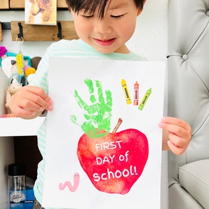 May include: A child is holding a white piece of paper with a green handprint, three crayons, a red apple with the text "FIRST DAY of school!" and a pink worm.