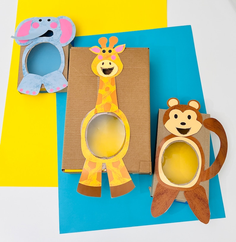 K&ouml;nnte beinhalten: Drei Pappkartons mit ausgeschnittenen Tiergesichtern. Die Kartons sind mit einer Giraffe, einem Elefanten und einem Affen verziert. Jeder Karton hat ein ausgeschnittenes Loch f&uuml;r ein Spiel.