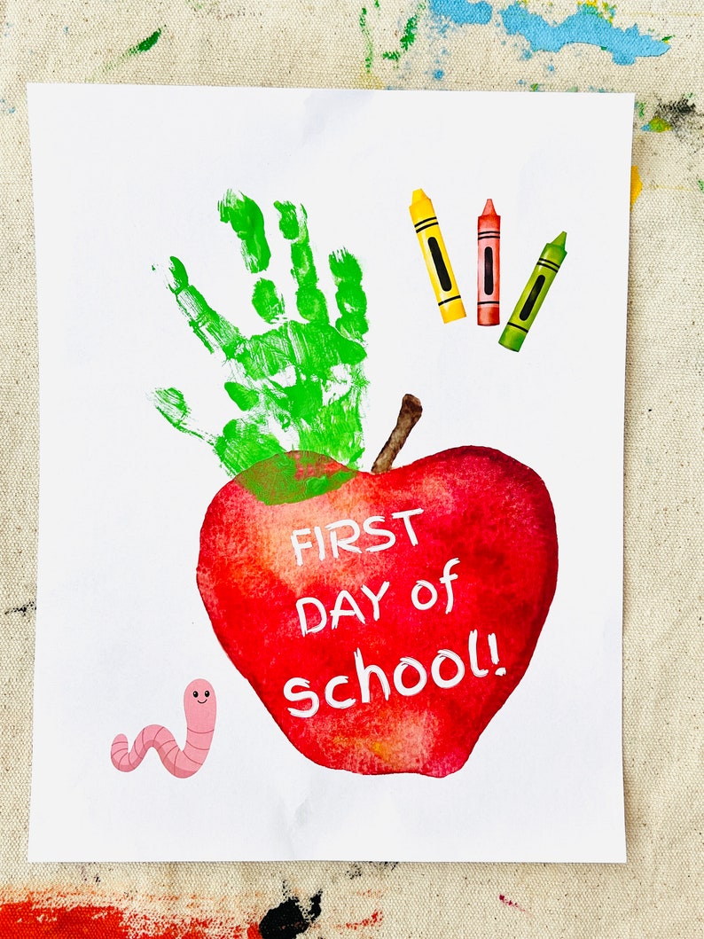 May include: A red apple with a green handprint on top and the words "FIRST DAY of school!" written on it. There are three crayons, one yellow, one red, and one green, and a pink worm on the white background.