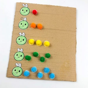 May include: A cardboard sheet with five green cartoon caterpillars with smiling faces. Each caterpillar has a corresponding number of pom-poms in a rainbow color pattern: red, orange, yellow, green, and blue.