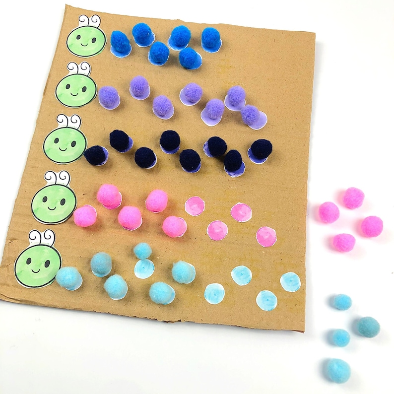 May include: A brown cardboard sheet with colorful pom-poms and painted dots. The pom-poms are arranged in rows, with each row containing a different color. There are also several green cartoon ladybugs with antennas and smiling faces.