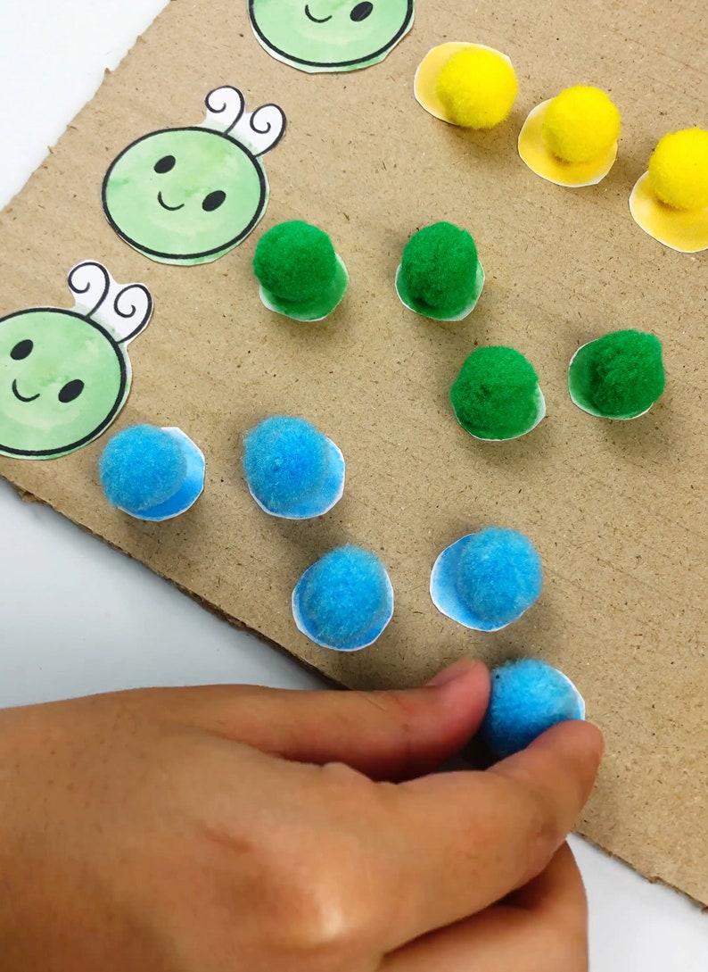 May include: A child's hand placing a blue pom-pom on a brown cardboard surface. There are green and yellow pom-poms already placed on the surface, along with cartoon images of green caterpillars.