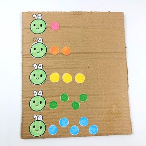 May include: A cardboard sheet with five rows of colorful dots and green cartoon faces. The first row has one pink dot, the second row has two orange dots, the third row has three yellow dots, the fourth row has four green dots, and the fifth row has five blue dots.