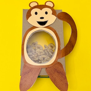 K&ouml;nnte beinhalten: Eine braune Pappbox in Form eines Affen mit einem runden Ausschnitt in der Mitte. Der Ausschnitt ist mit getrockneten Bananenscheiben gef&uuml;llt.
