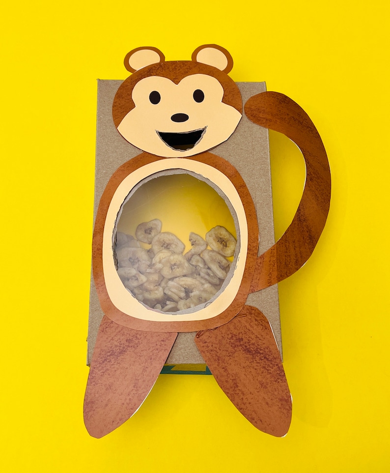 K&ouml;nnte beinhalten: Eine braune Pappbox in Form eines Affen mit einem runden Ausschnitt in der Mitte. Der Ausschnitt ist mit getrockneten Bananenscheiben gef&uuml;llt.