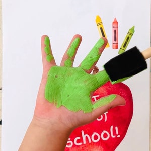 May include: A child's hand painted green is making a handprint on a red apple with the words "First Day of School!" written on it.  There are three crayons in the background.