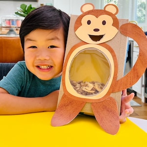 K&ouml;nnte beinhalten: Eine Kartonbox in Form eines Affen mit einem ausgeschnittenen Fenster, das mit M&uuml;sli gef&uuml;llt ist. Ein kleines Kind h&auml;lt die Box und l&auml;chelt.