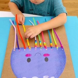 May include: A child is threading colorful pipe cleaners through holes in a cardboard cutout of a purple jellyfish with a smiling face. The jellyfish has white spots and pink cheeks. The pipe cleaners are threaded with colorful beads.