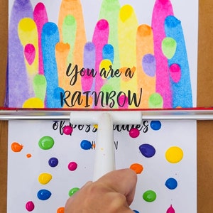 May include: A white card with a rainbow design and the text "You are a RAINBOW of good vibes". The card is being decorated with colorful paint dots.