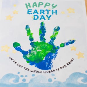 以下が含まれることがあります： 白いカードに、「Happy Earth Day」という緑と青の文字が書かれています。カードの中央には青と緑の手形があり、その周りに「We've got the whole world in our hands」という文字が書かれています。カードには黄色の星と青い雲があります。