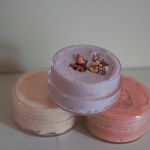 Puede incluir: Tres pequeños frascos de vidrio con tapas que contienen cremas de diferentes colores. El frasco superior contiene una crema de color púrpura claro con pétalos de rosa secos en la parte superior.