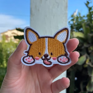 May include: Embroidered patch featuring a cartoon corgi dog with a happy expression. The patch has a white border, brown and tan fur, white ears, and pink details on the paws and tongue. The patch is held in a hand.
