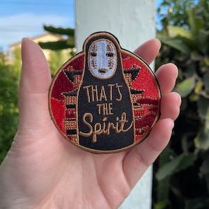 May include: Circular embroidered patch featuring a character from Spirited Away with a white face and black robe. The patch has a red border with a Japanese-style building design and the text "THAT'S THE Spirit" in gold lettering.
