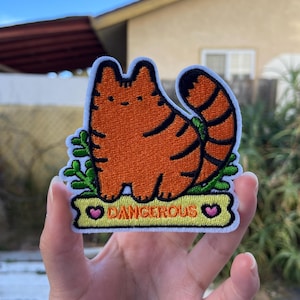 May include: Embroidered patch featuring an orange cartoon cat with black stripes and a curled tail. The patch is on a green leafy base with a yellow banner that reads "DANGEROUS" with pink hearts on each end.