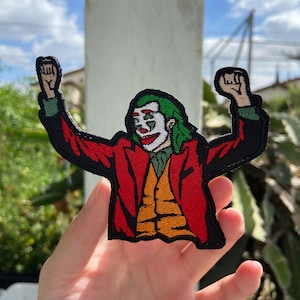 May include: Embroidered patch featuring the Joker character with green hair, white face paint, and a red suit. The patch has a black border and depicts the Joker with arms raised in a celebratory pose. The background shows a blue sky and greenery.