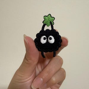 May include: A black embroidered patch featuring a cartoon-like creature with large white eyes and a green star-shaped element on top. The patch has a fluffy texture and small black legs.