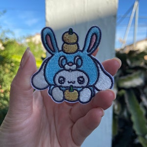 May include: Embroidered patch featuring a cartoon bunny wearing a blue hat with a pumpkin on top. The bunny has a white face, pink ears, and is holding a small pumpkin. The patch is outlined in white and is held in a hand.