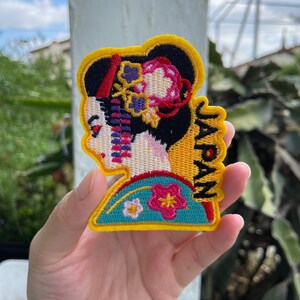 May include: Embroidered patch featuring a geisha in profile, with a black hairstyle adorned with floral accents. The patch has a yellow border and the word "JAPAN" in yellow. The geisha's kimono is teal with pink and white floral details.