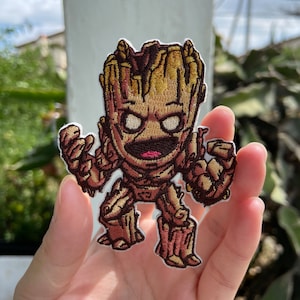 May include: Embroidered patch featuring a stylized Groot character from Guardians of the Galaxy. The patch has a brown and tan color scheme with detailed stitching, depicting Groot's wooden texture. The patch is held in a hand against a blurred background.