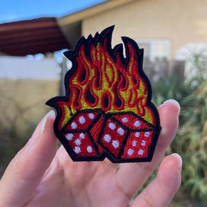 May include: Embroidered patch featuring two red dice with white dots, engulfed in flames. The flames are in shades of red and yellow, outlined in black. This patch is designed for application on clothing or accessories.