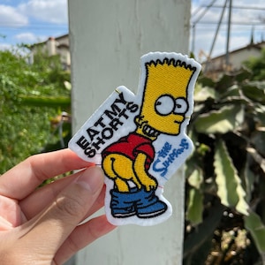 May include: Embroidered patch featuring Bart Simpson with the text "EAT MY SHORTS" and "The Simpsons". The patch shows Bart with his pants down, wearing a red shirt, blue jeans, and yellow skin. The patch has a white border.