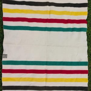 May include: A white blanket with horizontal stripes in black, yellow, red, and green. The blanket has a simple design and is made of a soft material. The blanket is folded and laid out on a green surface.