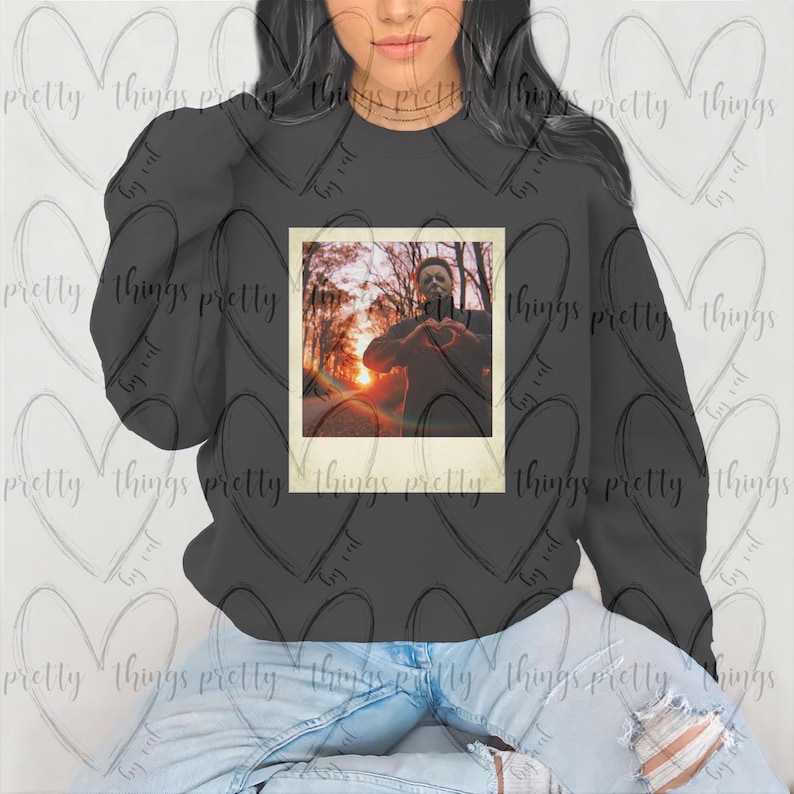 May include: A black sweatshirt with a vintage-style photo of a man in a horror movie mask. The photo is in a white frame with a light brown border. The man is wearing a dark jacket and is making a heart shape with his hands.