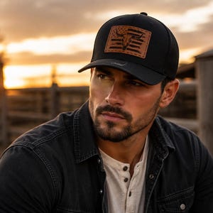 May include: A black baseball cap featuring a rectangular brown leather patch on the front. The patch displays a graphic of a bull skull centered over a stylized American flag design with horizontal stripes and stars. The cap has a curved brim.