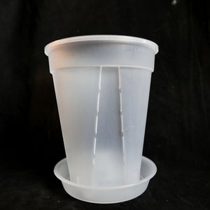 Może przedstawiać: Przezroczysta plastikowa doniczka z pasującą podstawką. Doniczka jest cylindryczna z pionowymi rowkami i szerokim kołnierzem. Podstawka jest okrągła i płytka. Przedmioty wykonane są z matowego, półprzezroczystego tworzywa sztucznego.