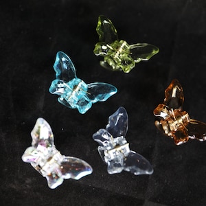 May include: Five colorful butterfly-shaped hair clips in shades of blue, green, orange, and clear. Each clip has a metal clasp and is made of translucent plastic, creating a shimmering effect against a black background.
