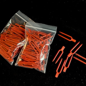 May include: Two clear plastic bags filled with a large quantity of small, red plastic tuning forks.  Five additional tuning forks are scattered on a black surface.