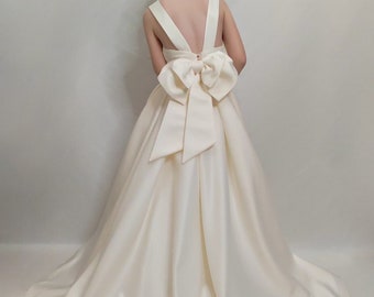 May include: A white satin dress with a large bow in the back. The dress has a sleeveless design and a flowing skirt.