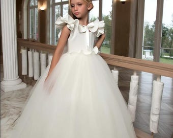 May include: A young person wearing a white formal dress with a full tulle skirt and ruffled cap sleeves. The dress has a satin bodice and a bow detail at the waist. The background features a grand staircase with arched windows and a chandelier.