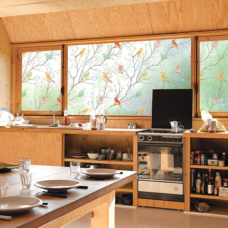 May include: A kitchen window with a frosted glass film featuring a colorful bird and branch design. The window is in a modern kitchen with wooden cabinets and a stove.