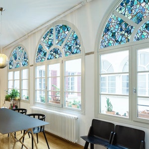 May include: Three arched windows with stained glass designs in a light blue and white color scheme. The windows are in a room with white walls and a wooden floor. There is a white radiator under the windows and two black chairs in the foreground.
