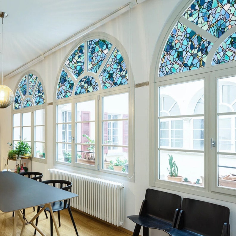May include: Three arched windows with stained glass designs in a light blue and white color scheme. The windows are in a room with white walls and a wooden floor. There is a white radiator under the windows and two black chairs in the foreground.