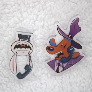May include: Two cartoon character acrylic charms. The first charm is a white rabbit with a black phone cord wrapped around its head and a black phone receiver hanging from its neck. The second charm is a brown dog wearing a purple suit, a blue tie, a purple hat, and holding a blue phone receiver to its ear.