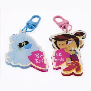 Puede incluir: Dos llaveros de mejores amigos, uno con un fantasma azul sosteniendo un corazón rosa que dice "Best" y el otro con una niña de cabello castaño sosteniendo un corazón rosa que dice "Friends".