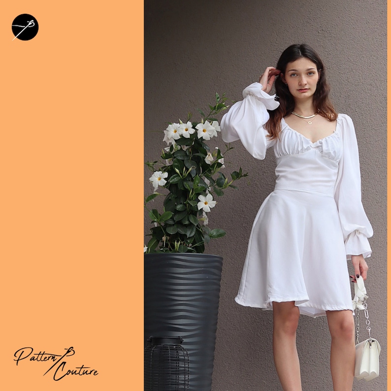 May include: A white dress with puffed sleeves and a sweetheart neckline. The dress has a fitted bodice and a flowing skirt. The model is standing in front of a wall with a potted plant.