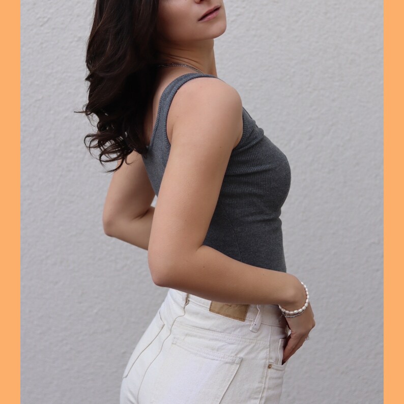 May include: A gray ribbed tank top and white jeans. The tank top has a scoop neck and a fitted silhouette. The jeans are high-waisted with a straight leg. A pearl bracelet is worn on the wrist.