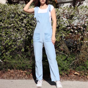 May include: A light blue overall jumpsuit with thin straps that tie at the shoulders. The jumpsuit has two large pockets on the front and a relaxed fit.