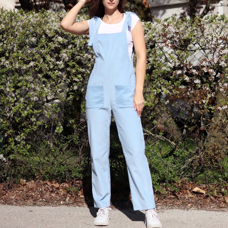 May include: A light blue overall jumpsuit with thin straps that tie at the shoulders. The jumpsuit has two large pockets on the front and a relaxed fit.