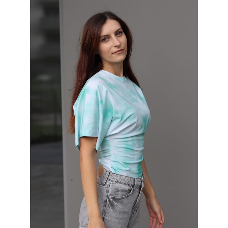 May include: A light blue and white tie-dye top with short sleeves. The top is ruched at the waist and has a round neckline. The model is wearing gray jeans. The background is a gray wall.